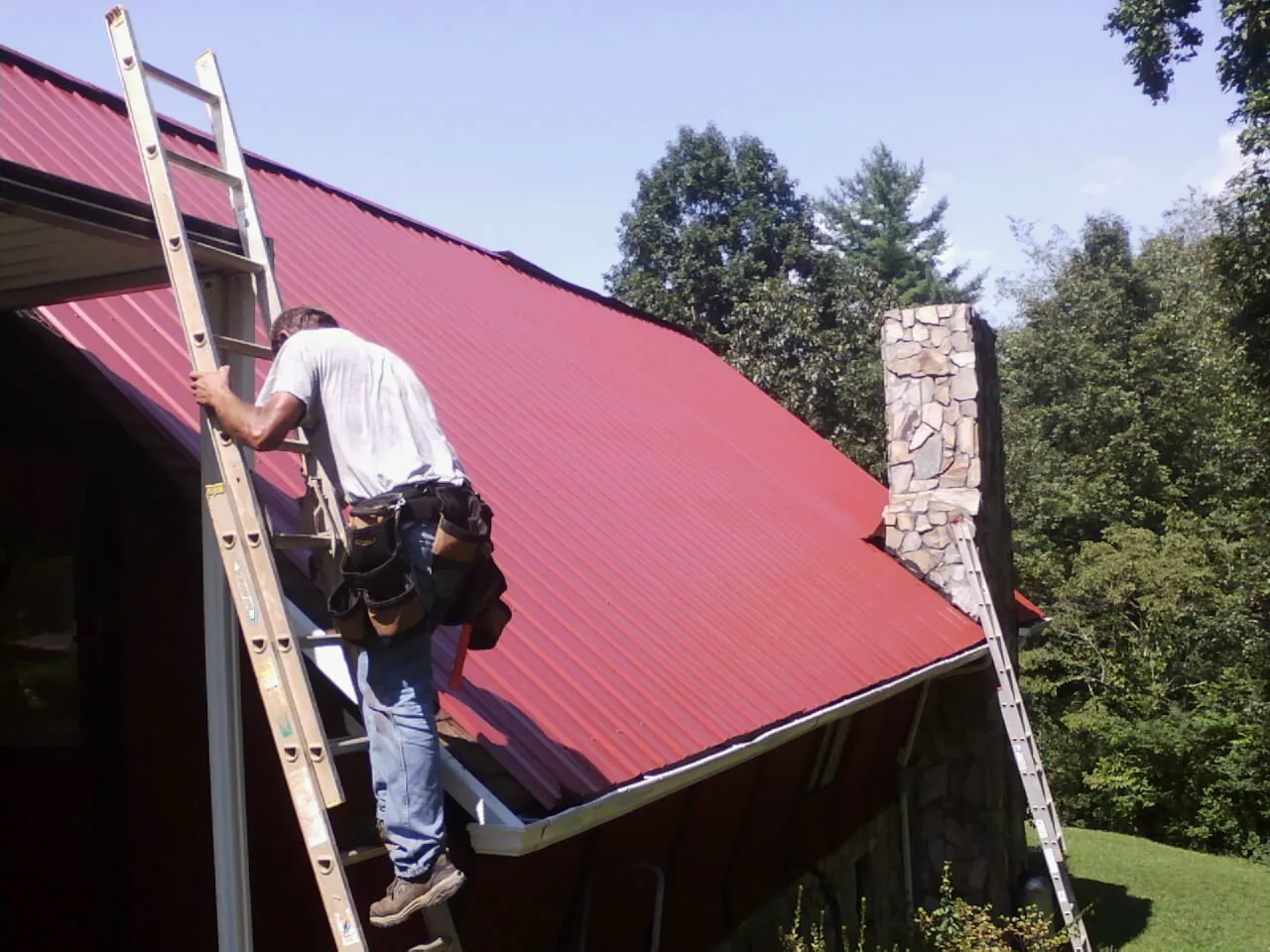 Roofer on a ladder beside a red metal roof and stone chimney — Soffit & Fascia Repair by Roofing Brothers in Vancleave